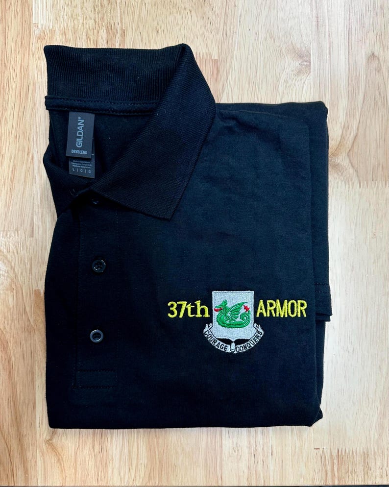 May include: A black polo shirt with a collar and three buttons. The chest features an embroidered emblem with a green dragon on a shield, flanked by the yellow text 37th and ARMOR, with a banner below reading COURAGE CONQUERS.