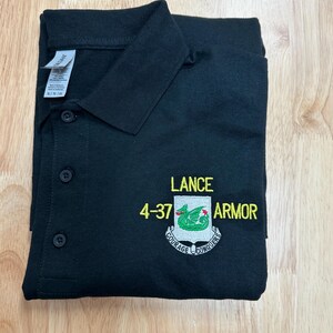 May include: A black polo shirt with a collar and three buttons. The chest features an embroidered crest with a green dragon, the text LANCE 4-37 ARMOR, and a banner below the shield that reads COURAGE CONQUERS in white and yellow thread.
