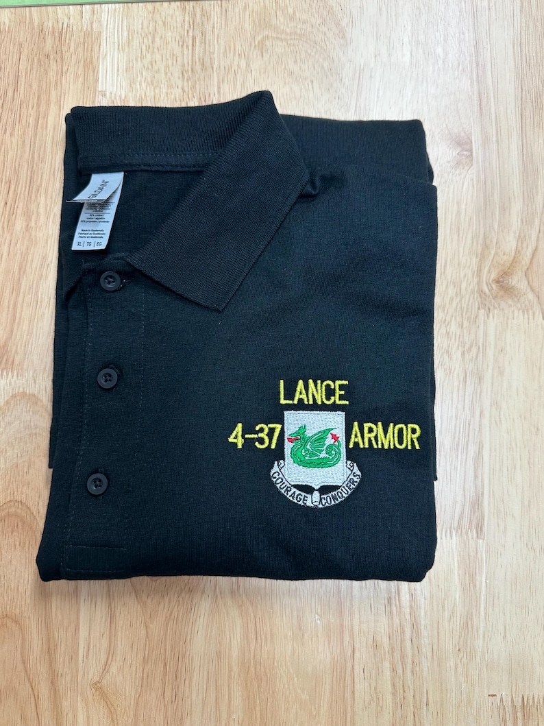 May include: A black polo shirt with a collar and three buttons. The chest features an embroidered crest with a green dragon, the text LANCE 4-37 ARMOR, and a banner below the shield that reads COURAGE CONQUERS in white and yellow thread.