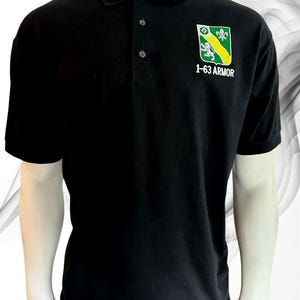 May include: A black short-sleeved polo shirt featuring a three-button placket and a collar. On the left chest, there is an embroidered shield-shaped emblem in green, yellow, and white, displaying a lion and the text 1-63 ARMOR below it.