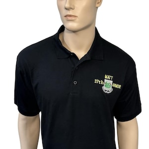 May include: A black short-sleeved polo shirt with a collar and a three-button placket. Embroidered on the left chest in yellow and green thread is the text MATT 37th ARMOR above a shield-shaped emblem featuring a green dragon graphic.