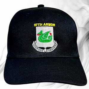 May include: A black baseball cap featuring an embroidered emblem on the front. The emblem displays a green dragon on a white shield, topped with the yellow text 37TH ARMOR and a white banner below with the black text COURAGE CONQUERS.