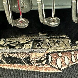 May include: A black fabric surface featuring an embroidered design of a military tank in shades of tan, brown, and black. Above the embroidery, several metal embroidery machine needles and presser feet are positioned in a row.