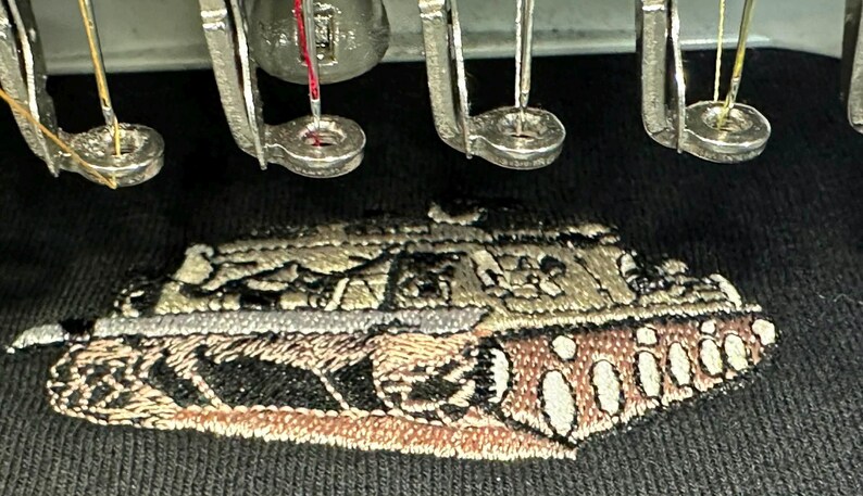 May include: A black fabric surface featuring an embroidered design of a military tank in shades of tan, brown, and black. Above the embroidery, several metal embroidery machine needles and presser feet are positioned in a row.