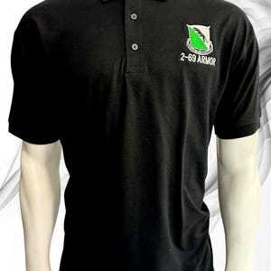 May include: A black short-sleeved polo shirt featuring a three-button placket and a collar. On the left chest, there is an embroidered emblem with a green and white shield, a black panther, and the text 2-69 ARMOR and VITESSE ET PUISSANCE.
