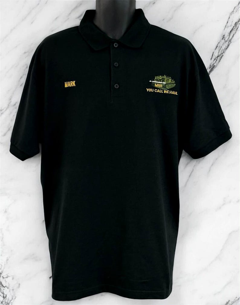 May include: A black short-sleeved polo shirt with a collar and three buttons. The right chest features the name MARK in yellow embroidery. The left chest displays an embroidered green tank with the text M88 YOU CALL WE HAUL in yellow.
