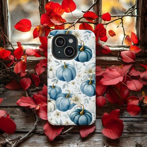 Könnte beinhalten: Smartphone-Hülle mit einem sich wiederholenden Muster aus blauen Kürbissen mit goldenen Stielen und weißen Blumen auf weißem Hintergrund. Die Hülle hat ein schwarzes Kameragehäuse. Rote Blätter und eine rustikale Holzoberfläche umgeben die Hülle.