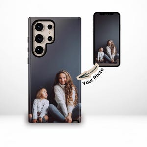 Puede incluir: Funda de teléfono inteligente negra con una fotografía de dos personas. La funda tiene un acabado brillante y un recorte para la cámara. Un segundo teléfono muestra la misma imagen. También se ven las palabras "Your Photo".
