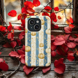 Könnte beinhalten: Eine Smartphone-Hülle mit herbstlichem Design. Sie zeigt einen blau-weiß gestreiften Hintergrund mit Kürbis- und Rankenillustrationen. Die Hülle ist von roten Blättern umgeben und liegt auf einer rustikalen Holzoberfläche.