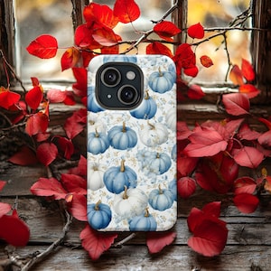 May include: A white and blue pumpkin-themed phone case. The case features a pattern of blue and white pumpkins with gold stems, set against a floral background. The phone case is displayed near red leaves and a wooden window.