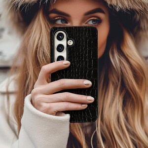 Op de afbeelding: Zwarte telefoonhoes met een textuurpatroon dat op krokodillenleer lijkt. De hoes heeft een uitsparing voor de camera met drie lenzen en een flitser. De telefoonhoes wordt voor het gezicht van een persoon gehouden.