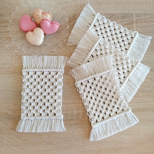 Puede incluir: Cuatro posavasos de crochet blanco con flecos, cada uno con un patrón cuadrado. Los posavasos están dispuestos en una pila, con el posavasos superior ligeramente desplazado de los demás.