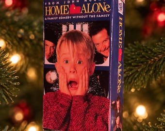 May include: A miniature replica of the Home Alone VHS tape box ornament featuring a red ribbon hanger. The front displays the movie title, the text From John Hughes, and a graphic of a child with hands on their face, flanked by two men.