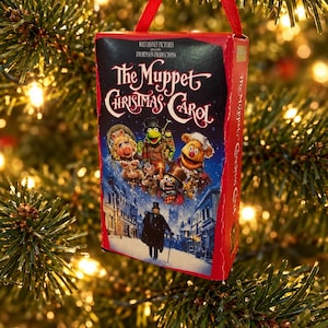 May include: A rectangular Christmas tree ornament shaped like a VHS tape case for The Muppet Christmas Carol. The front features the movie title, Walt Disney Pictures and Jim Henson Productions text, and illustrations of Muppet characters in a snowy scene.