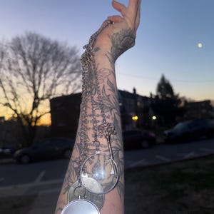 Puede incluir: Un collar plateado con múltiples colgantes, incluyendo una llave, una lupa y colgantes redondos. El collar cuelga de una cadena y se muestra sobre un fondo borroso al aire libre.