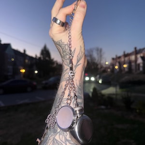 Puede incluir: Un llavero plateado con un pequeño frasco, un espejo compacto redondo y un dije de llave decorativa. Los objetos están suspendidos de una cadena, con un cielo crepuscular de fondo.