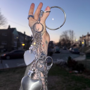 Puede incluir: Una colección de amuletos y colgantes plateados, que incluyen un frasco en forma de corazón, un relicario en forma de corazón y tijeras decorativas, unidos a un llavero. El llavero se sostiene contra un cielo crepuscular, con un gran anillo plateado en la parte superior.