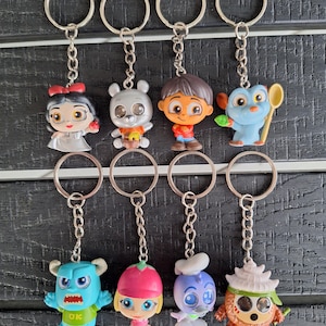 May include: A collection of eight character keychains. The keychains feature various cartoon characters in vibrant colors, each attached to a silver chain and ring. The characters include a princess, a bear, a boy, a blue bird, a monster, a girl, a chef, and a creature.
