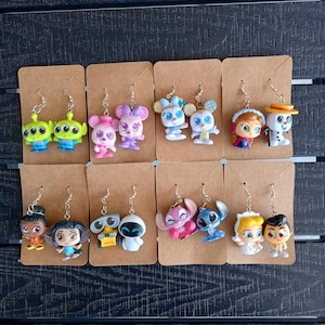 May include: A set of 10 pairs of dangle earrings featuring various Disney characters. The earrings are made of plastic and have a silver-toned metal hook. The characters include Stitch,  Elsa and Olaf,  Mike and Sully,  Wall-E and Eve,  Tinkerbell and  a couple from the movie Tangled.