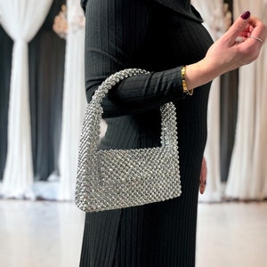 May include: A silver beaded handbag with a rectangular shape and a short handle. The bag is made of small, round beads that are tightly woven together.