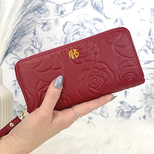 May include: A deep red, textured leather wallet featuring an embossed rose pattern and a gold-toned circular monogram with the letters RKS. The wallet includes a matching red wristlet strap attached to a gold-toned zipper pull and hardware.