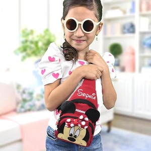 May include: A white short-sleeved shirt with pink hearts, blue jeans, and round cream-colored sunglasses. A red crossbody sling bag features the name Sophia in white script, the word Minnie, and a graphic of Minnie Mouse with a polka dot bow.