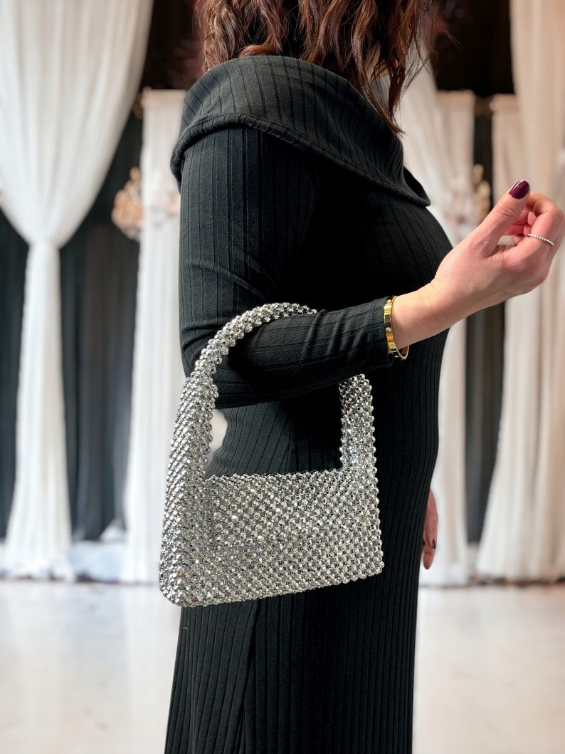 May include: A silver beaded handbag with a rectangular shape and a short handle. The bag is made of small, round beads that are tightly woven together.