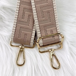 Pode incluir: Uma al&ccedil;a de bolsa tecida bege e creme com um padr&atilde;o geom&eacute;trico. A al&ccedil;a tem ferragens de metal dourado, incluindo fechos e an&eacute;is retangulares. A al&ccedil;a &eacute; exibida em uma superf&iacute;cie branca e fofa.
