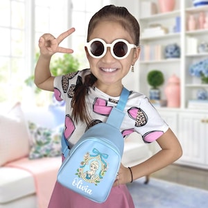 Puede incluir: Una mochila bandolera azul claro con un diseño floral y el nombre "Olivia". La mochila tiene una correa azul y un cierre de cremallera. La niña lleva una camiseta blanca con corazones rosas y estampado de leopardo.