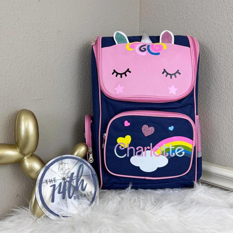 Op de afbeelding: Een marineblauwe rugzak met een roze eenhoorngezicht op de bovenflap, inclusief gesloten ogen, oren en een hoorn. Het voorvak toont een regenboog, een witte wolk, een glitterend hart en de naam Charlotte in witte letters gedrukt.
