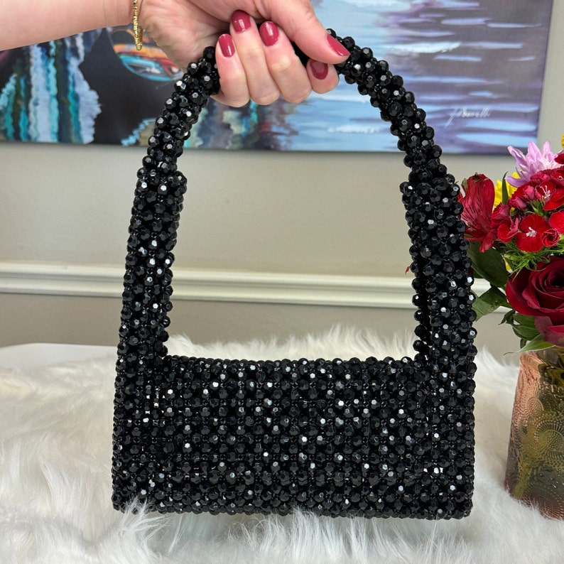 May include: A small black beaded handbag with a top handle. The bag is made of small, black beads that are sewn together to create a textured surface.