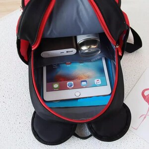 May include: A black backpack with red trim and a red zipper, shown from an open top-down perspective. Inside, the bag contains a silver tablet with a colorful screen, a dark rectangular power bank, and a small silver cylindrical container.