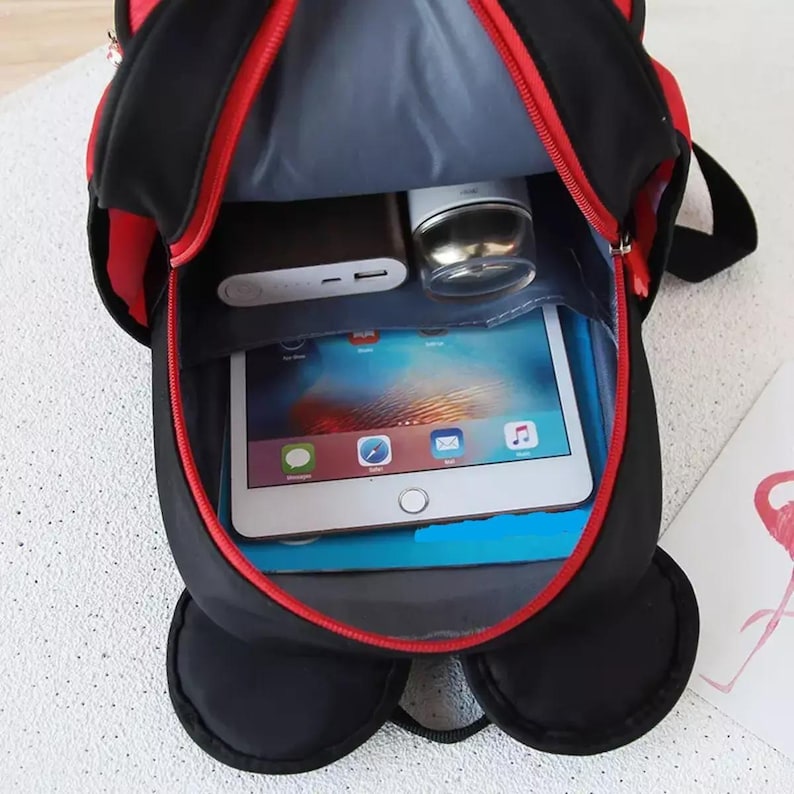 May include: A black backpack with red trim and a red zipper, shown from an open top-down perspective. Inside, the bag contains a silver tablet with a colorful screen, a dark rectangular power bank, and a small silver cylindrical container.