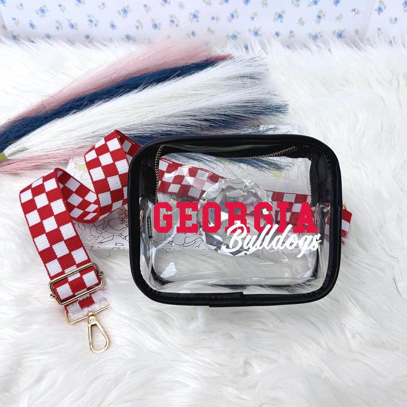 May include: A clear crossbody bag with a black trim and a red and white checkered strap. The bag has the words "GEORGIA Bulldogs" printed on the front in red and white.
