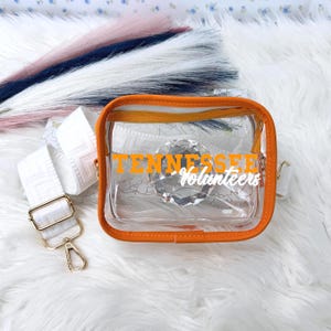May include: Clear crossbody bag with orange trim and a white strap. The bag features the text "Tennessee Volunteers" in orange. The strap is white with a geometric pattern and gold-colored hardware.