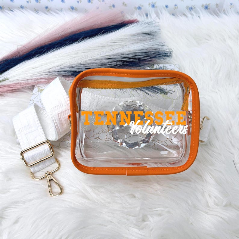 May include: Clear crossbody bag with orange trim and a white strap. The bag features the text "Tennessee Volunteers" in orange. The strap is white with a geometric pattern and gold-colored hardware.