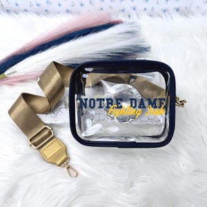 May include: A clear, rectangular Notre Dame Fighting Irish bag with a navy blue trim and a gold-colored adjustable strap. The bag features the text "Notre Dame Fighting Irish" in navy blue and gold lettering. The bag is on a white, fluffy surface.