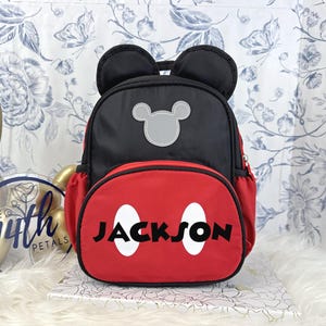 May include: A black and red backpack featuring two rounded ears at the top, a grey mouse-shaped silhouette patch, and a front pocket with two white oval shapes and the name JACKSON printed in black lettering across the center of the red section.