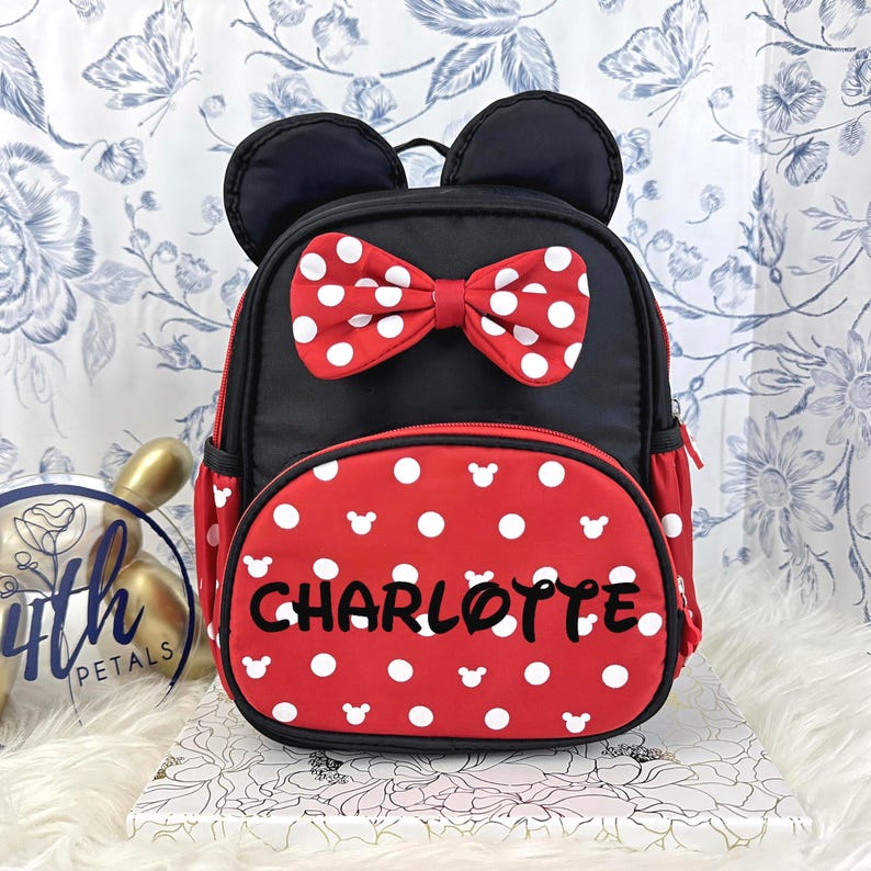 May include: A black and red backpack featuring a red bow with white polka dots, black mouse ears, and a red front pocket with white polka dots and mouse head silhouettes. The name CHARLOTTE is printed in black letters on the front pocket.