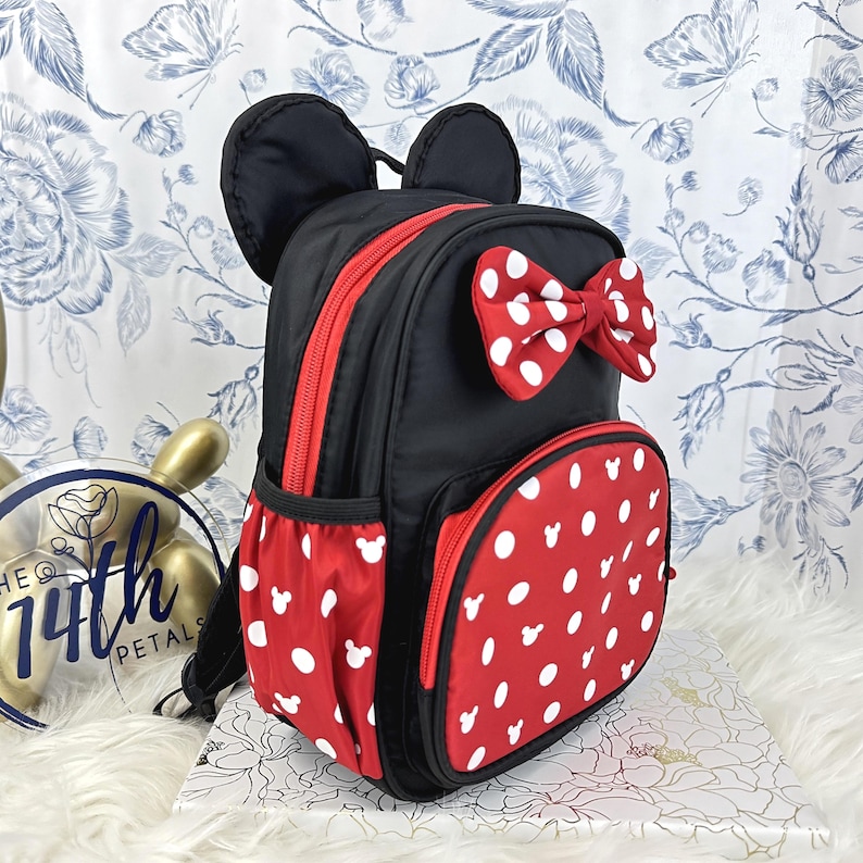May include: A black backpack featuring two rounded ears at the top, a red bow with white polka dots, and a front pocket with a red and white polka dot pattern that includes small white mouse head silhouettes. The bag has red zippers and side pockets.