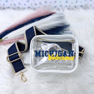 May include: A clear, square crossbody bag with a white trim and gold zipper. The bag features the text "MICHIGAN Wolverines" in blue and yellow. A navy blue and tan strap with gold hardware is attached.