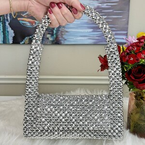 May include: A silver beaded handbag with a top handle. The bag is made of small, round beads that are tightly woven together.