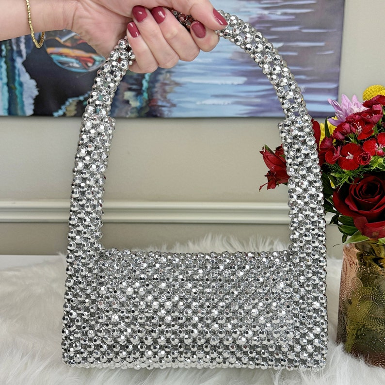 May include: A silver beaded handbag with a top handle. The bag is made of small, round beads that are tightly woven together.