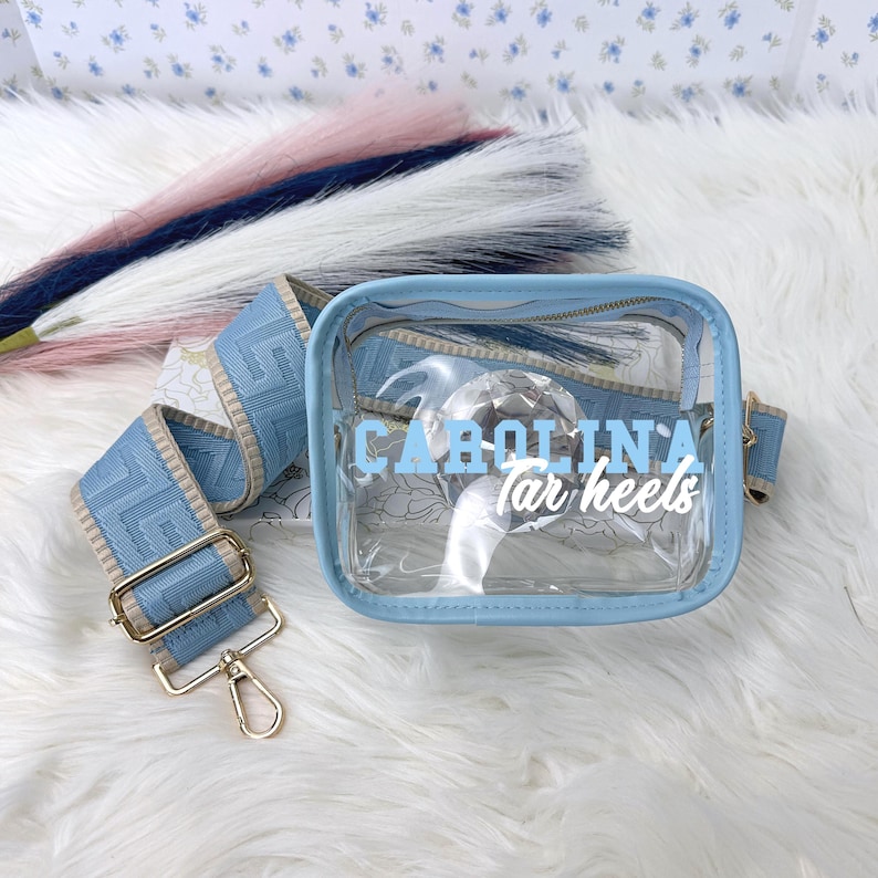 May include: A clear, light blue fanny pack with the text "Carolina Tar Heels" in white. The bag has a matching light blue strap with gold-colored hardware. The strap is patterned with a light blue design. The bag is on a white surface.