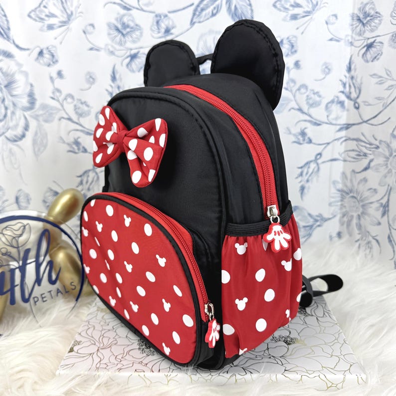May include: A black mini backpack featuring two rounded ears on top, a red front pocket with white polka dots and small white mouse head silhouettes, a red bow with white polka dots, and red side pockets with white polka dots and mouse head icons.