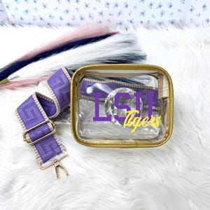 May include: A clear, rectangular cosmetic bag with a gold trim and a purple strap. The bag features the text "LSU Tigers" in purple and yellow. The strap is purple with a geometric design and gold hardware.