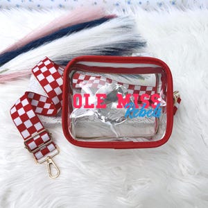 May include: A clear, rectangular crossbody bag with a red trim and zipper. The bag features a red and white checkered strap and the text "OLE MISS Rebels" in blue and red. The bag has a gold-colored clasp.
