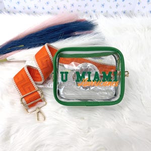 May include: Clear square crossbody bag with green trim and an orange strap. The strap has a repeating pattern. The bag has the text "U Miami Hurricanes" in green and orange. The bag has a gold-colored clasp.