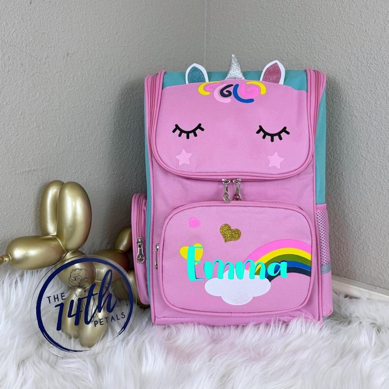 Op de afbeelding: Een roze rugzak met een eenhoorngezicht met gesloten ogen, een zilveren hoorn en oren. Het voorvak toont een regenboog, een witte wolk, een goudkleurig glitterhart en de naam Emma in turquoise schrift. De zijkanten zijn turquoise met gaasvakken.