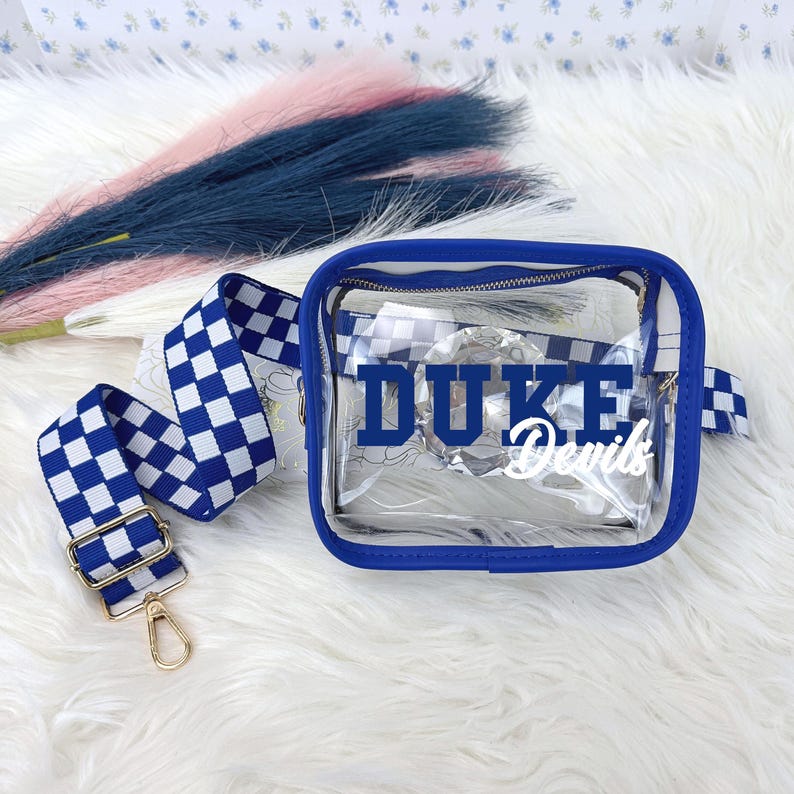May include: A clear, square crossbody bag with a blue trim and a blue and white checkered strap. The bag has the words "DUKE Devils" printed on the front in blue and white. The strap has gold-colored hardware.